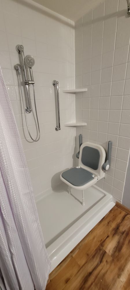White accessible shower with seat, grab bar, and shower head, in a bathroom with wood flooring.