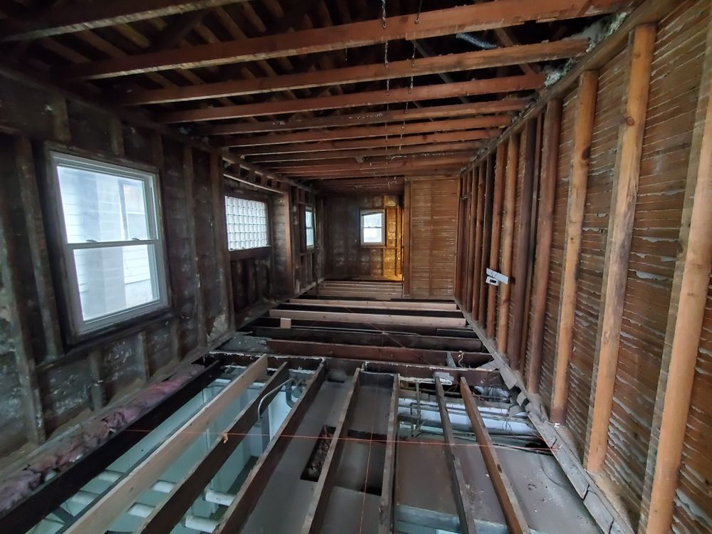 Interior of a room undergoing renovation; exposed beams, studs, and floor joists; windows along the wall.