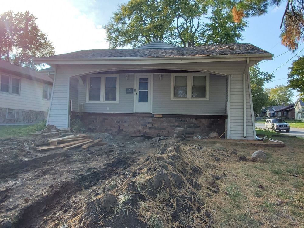 House with porch; dirt mounds in front. Gray siding, brown stone foundation, blue door.