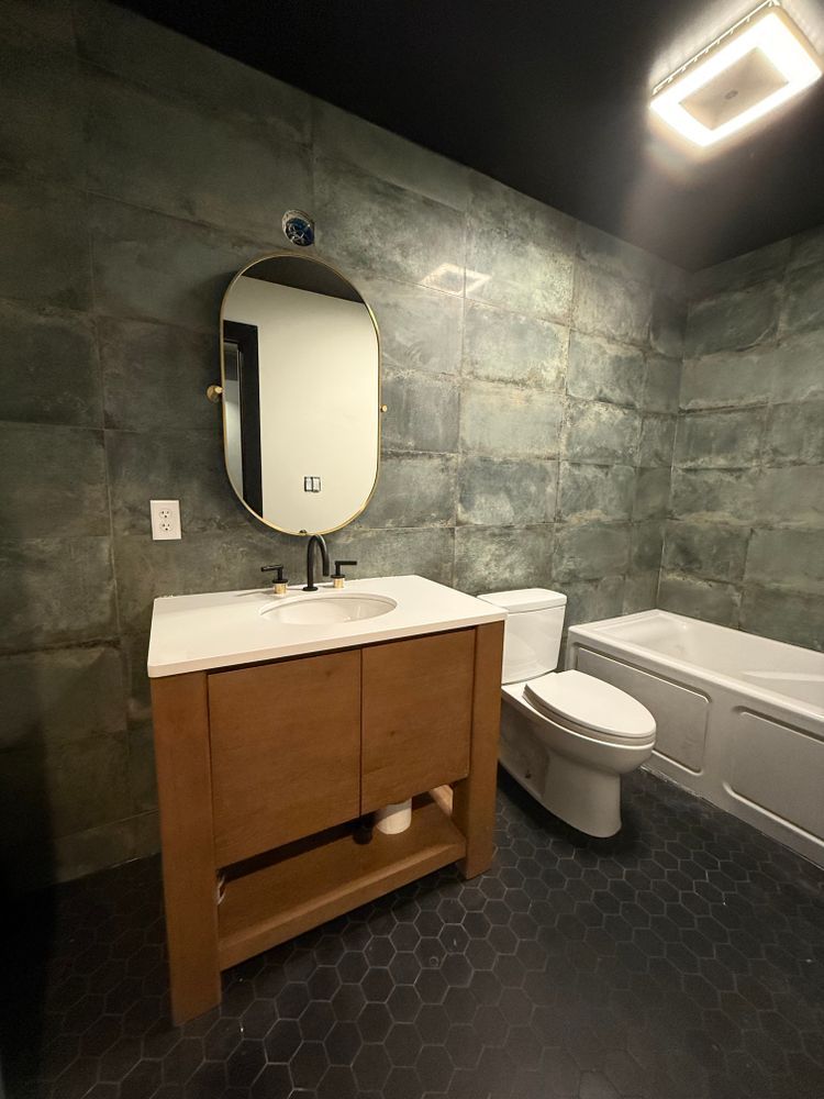 Bathroom with wooden vanity, oval mirror, toilet, and bathtub; walls are green and floor is dark hexagon tiles.