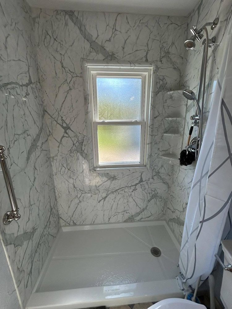 Bathroom with marble-patterned shower walls, window, showerhead, and grab bar.