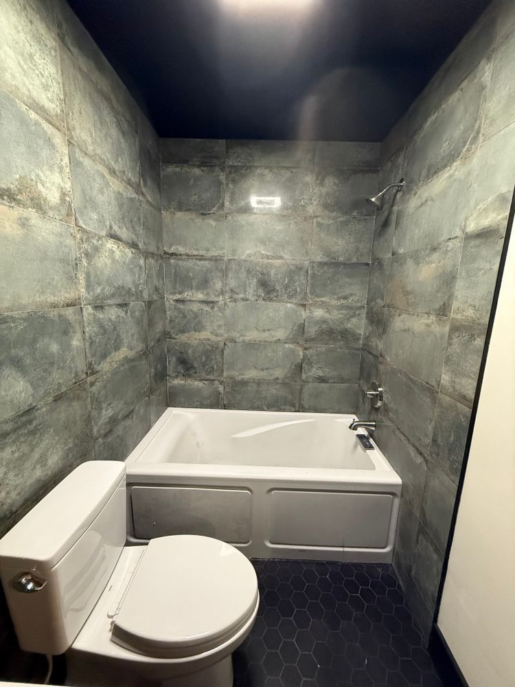 Bathroom with gray tile walls, a white tub and toilet, and a dark tiled floor.