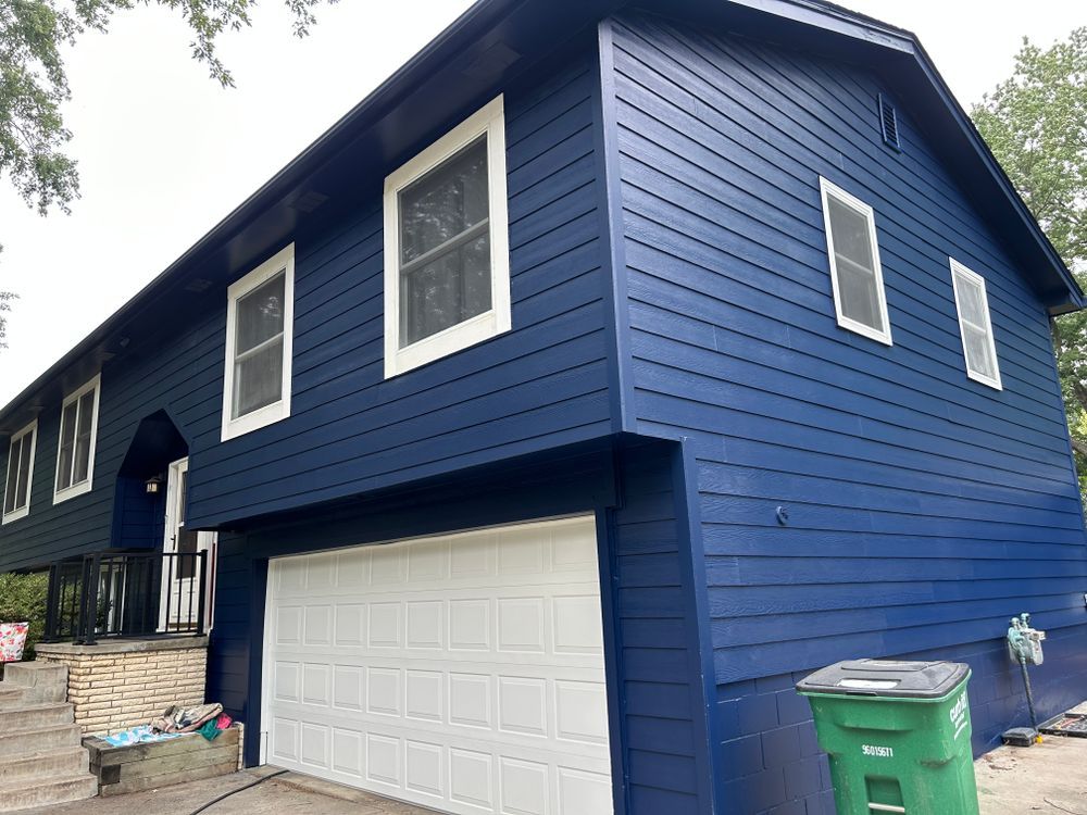 Blue house with white trim and garage door; green trash bin in front.