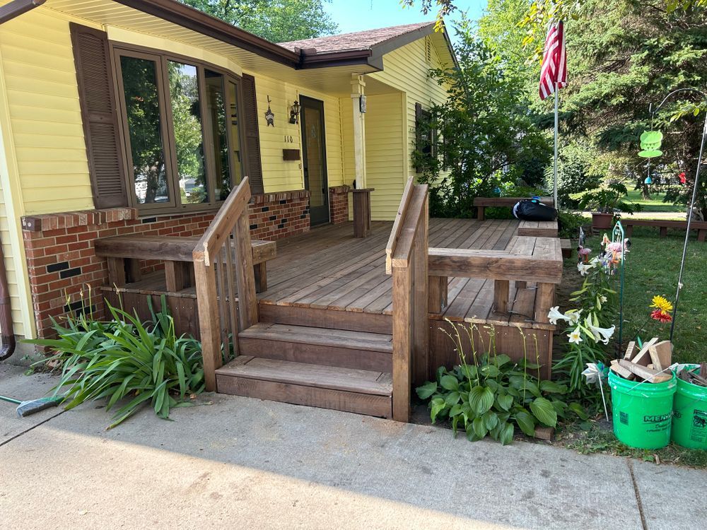 Yellow house with brown porch, steps, and wooden railing. American flag flies. Green plants and buckets in yard.