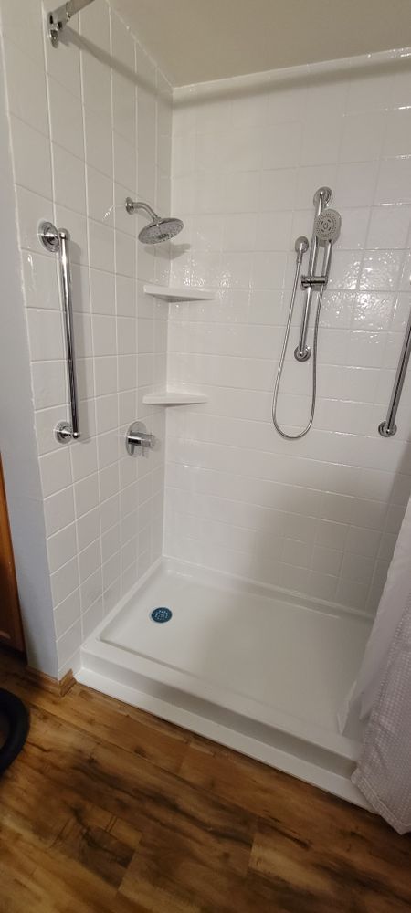 A white shower stall with grab bars, shower head, and handheld shower on a wood floor.
