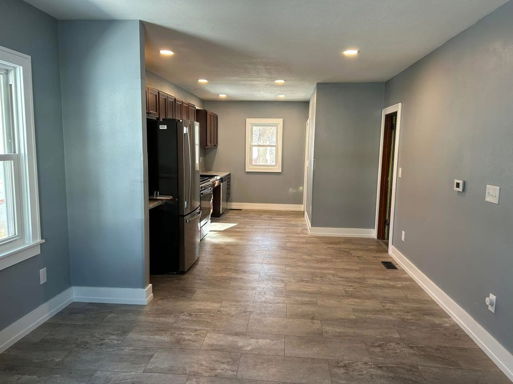 Empty living room with gray walls and wood-look flooring, leading into a kitchen with dark appliances.