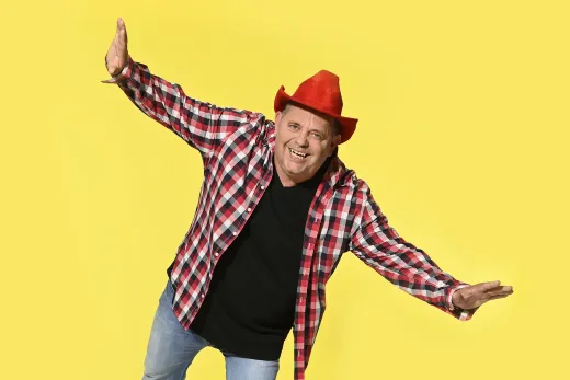 Ein Mann mit einem roten Cowboyhut und einem karierten Hemd steht mit ausgestreckten Armen da.