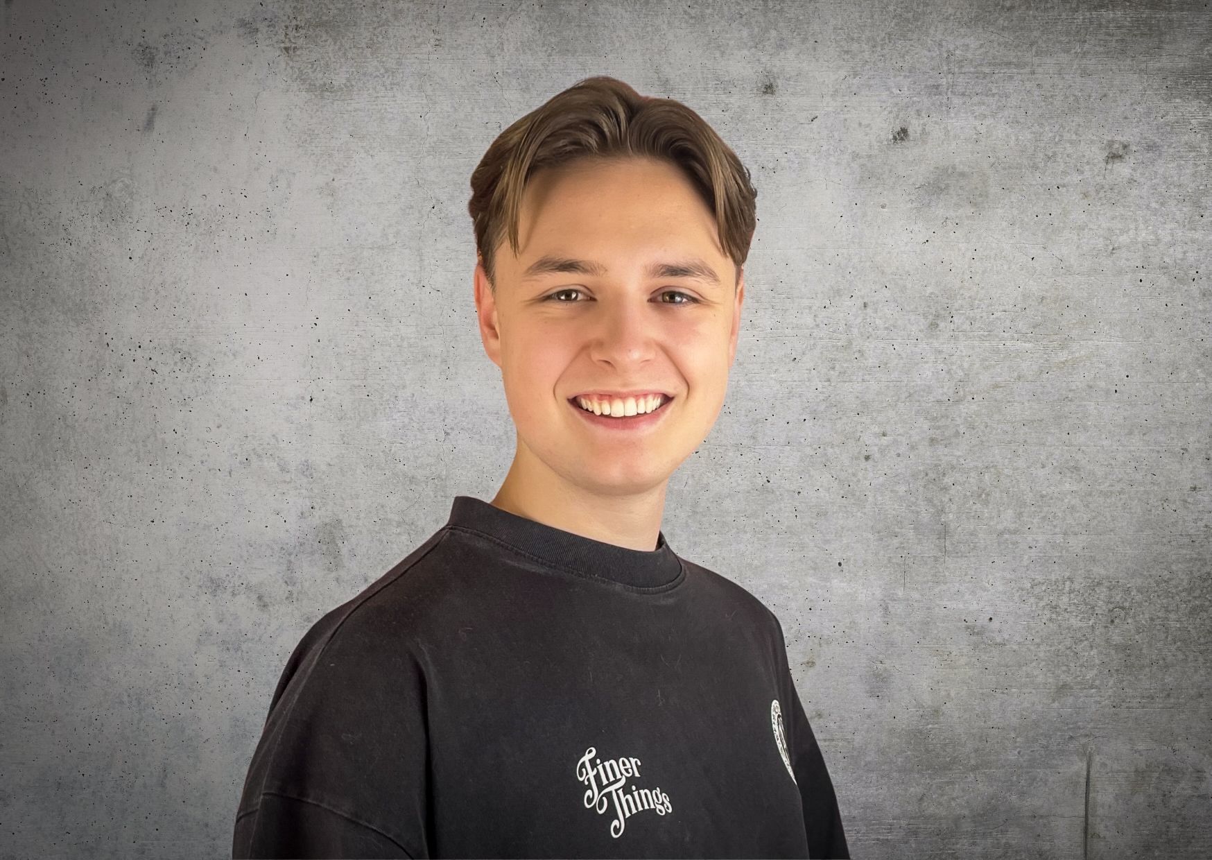 Ein junger Mann in einem schwarzen Sweatshirt lächelt vor einer Betonwand.