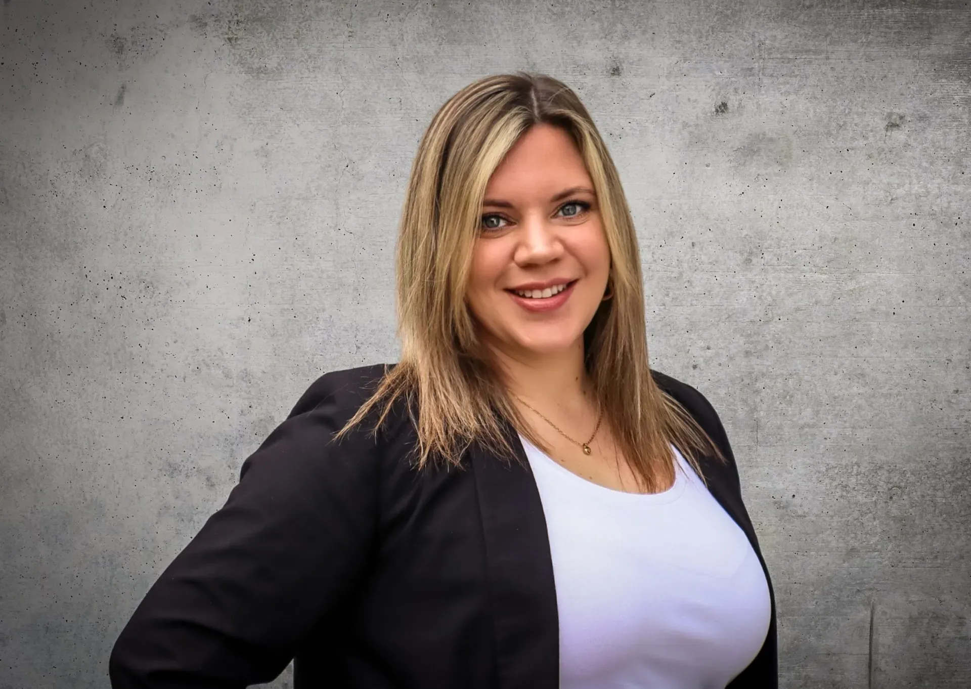 Eine Frau in einer schwarzen Jacke und einem weißen Hemd steht vor einer Betonmauer.