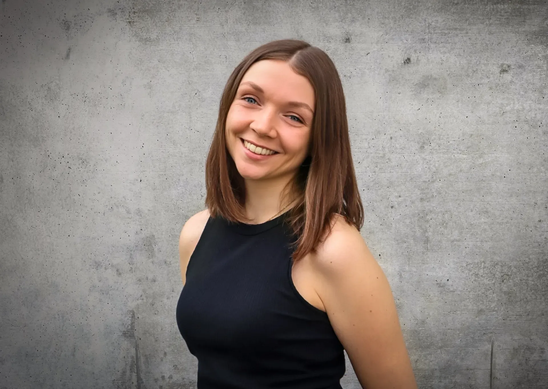 Eine Frau in einem schwarzen Tanktop lächelt vor einer Betonwand.