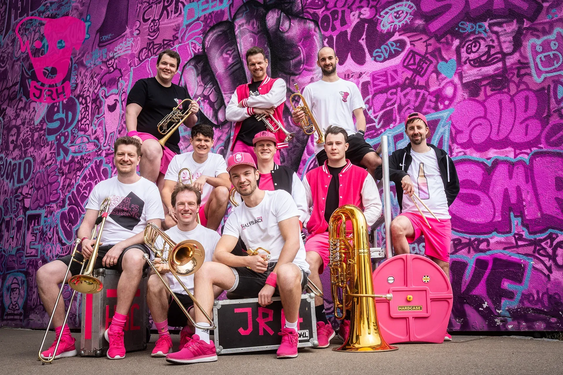 Eine Gruppe Männer posiert mit ihren Instrumenten vor einer rosa Wand für ein Foto.