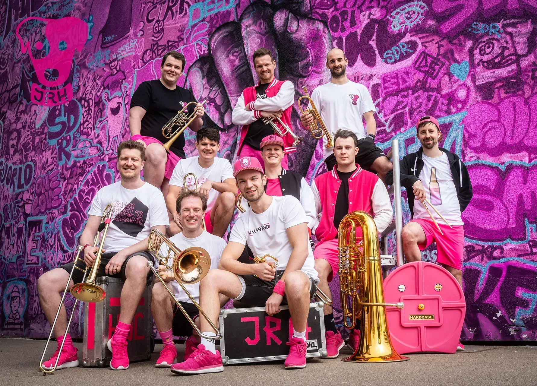 Eine Gruppe Männer posieren mit ihren Instrumenten vor einer Pinken Wand.