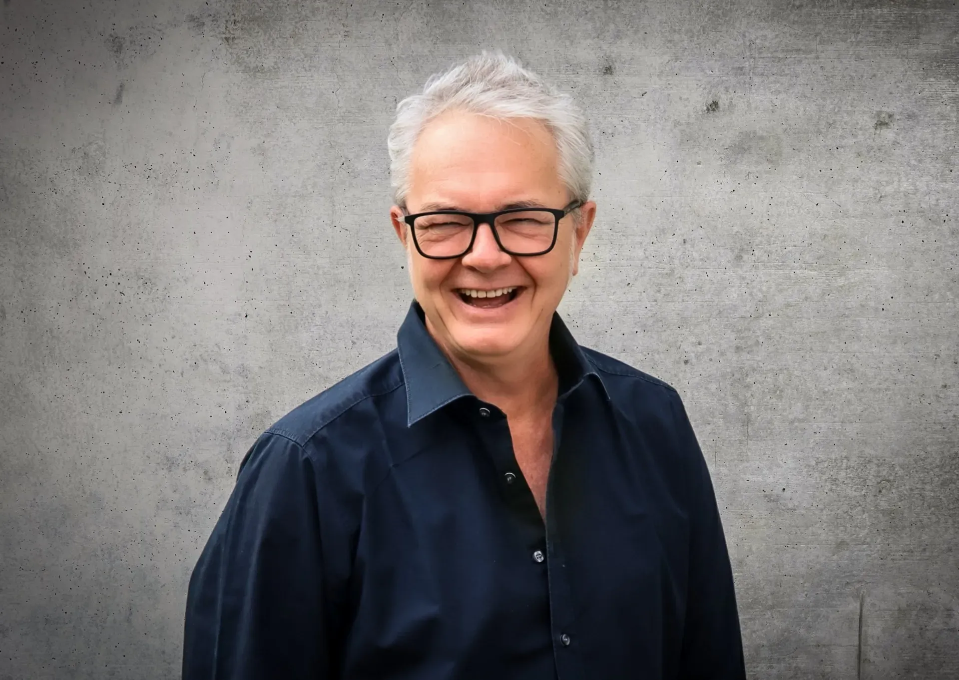 Ein Mann mit Brille und blauem Hemd lächelt vor einer Betonwand.