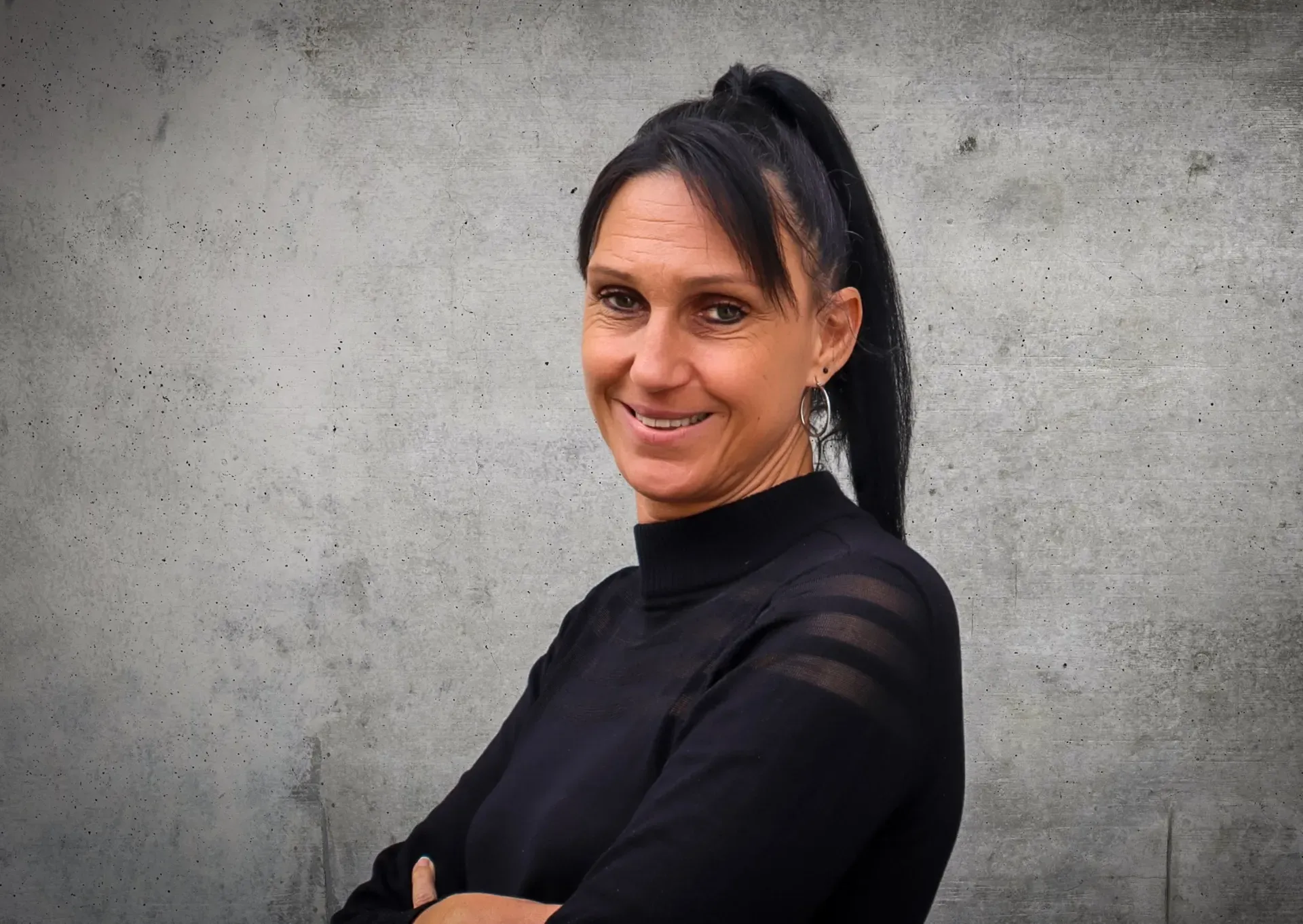 Eine Frau in einem schwarzen Hemd lächelt und hat die Arme verschränkt.