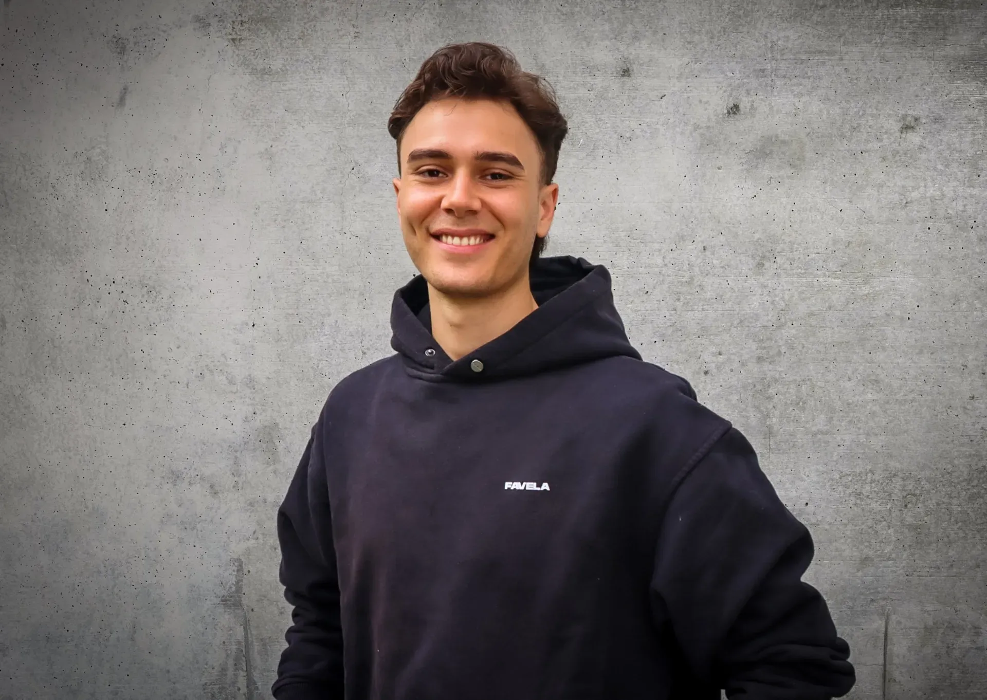 Ein junger Mann mit einem schwarzen Kapuzenpullover lächelt in die Kamera.