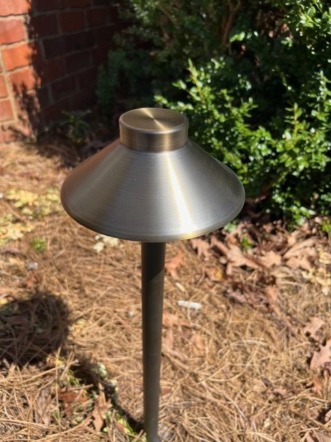 A stainless steel light is sitting in the dirt in front of a brick wall.