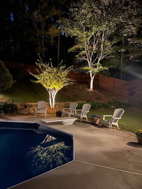 A patio with chairs and trees next to a pool at night.