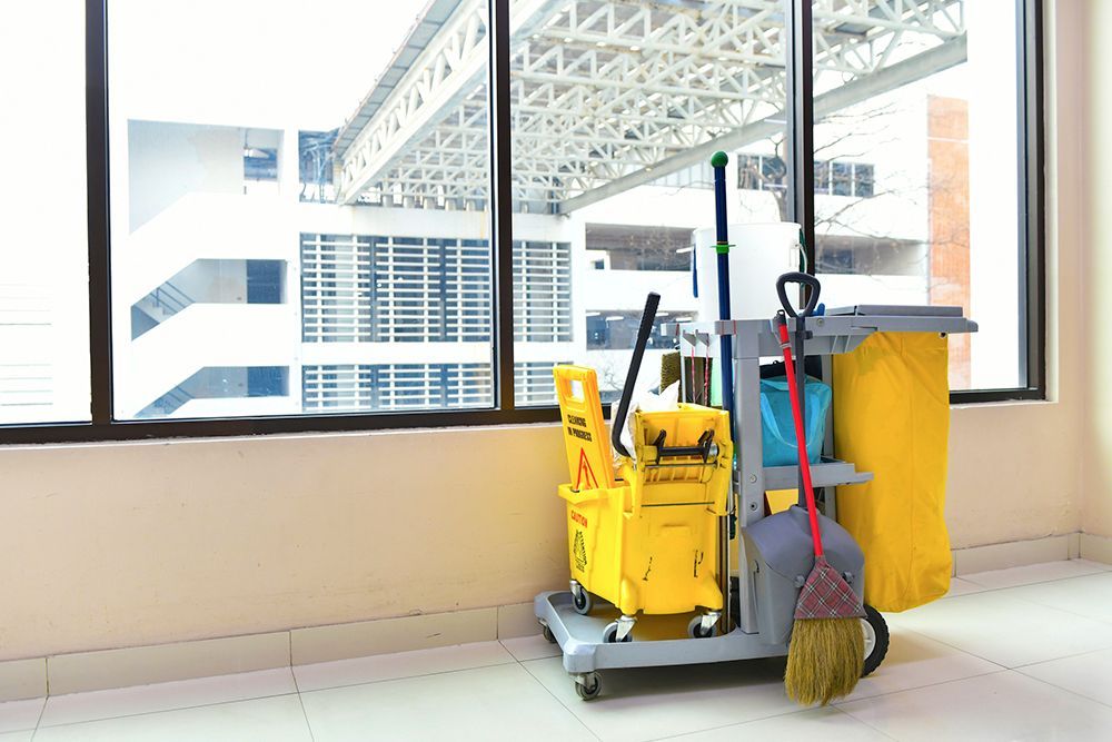 A cleaning cart with a mop , bucket , and broom in a room next to a window.