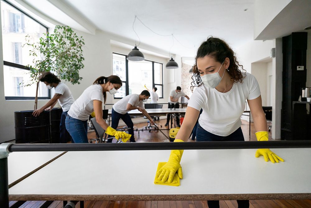 A group of people wearing masks and gloves are cleaning tables in an office.