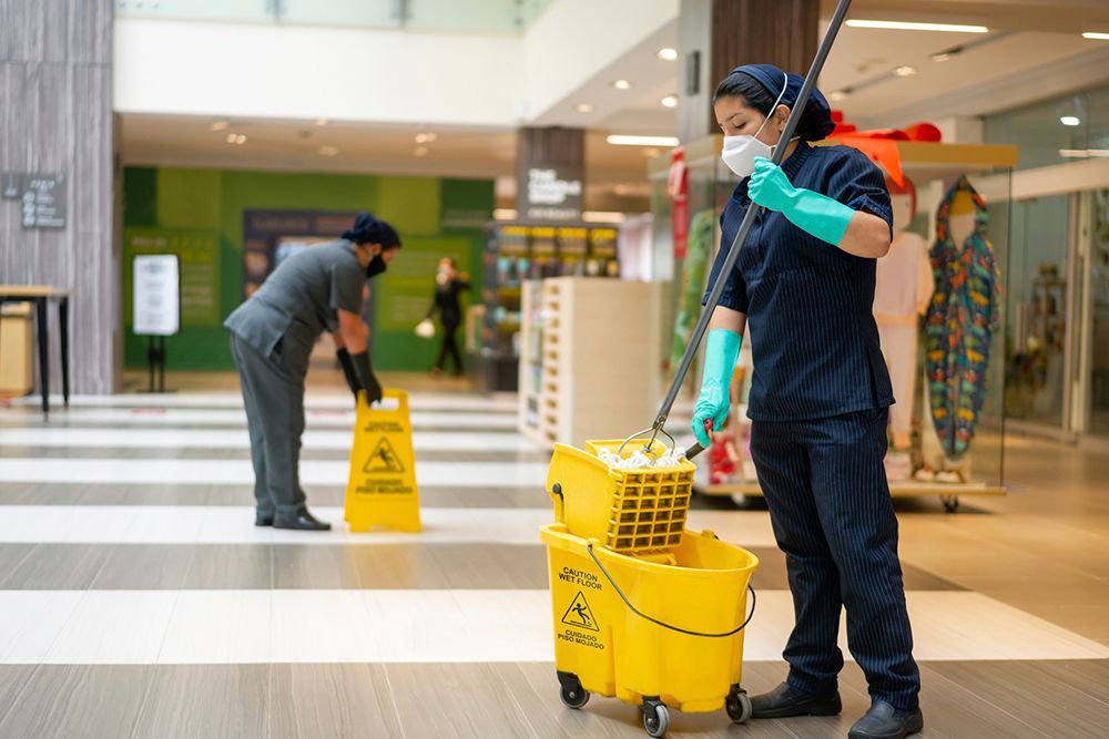 A woman wearing a mask and gloves is mopping the floor of a mall. A woman wearing a mask and gloves is mopping the floor of a mall.