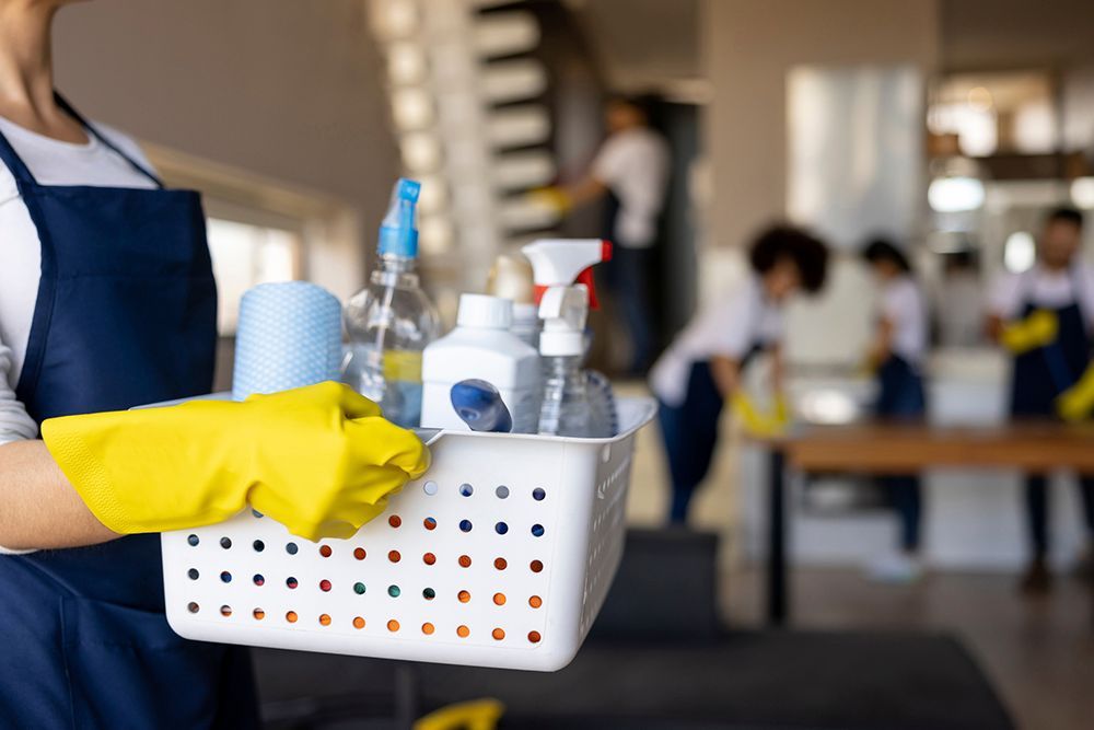 A woman in yellow gloves is holding a basket of cleaning supplies.