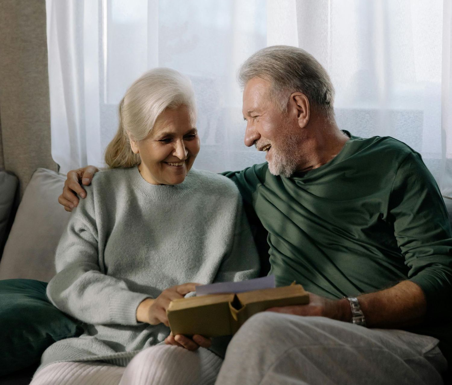 An elderly couple is sitting on a couch holding a gift box.