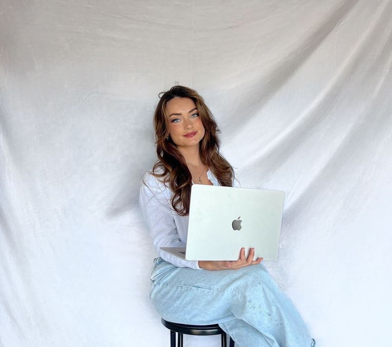 A woman is sitting on a stool holding an apple laptop.