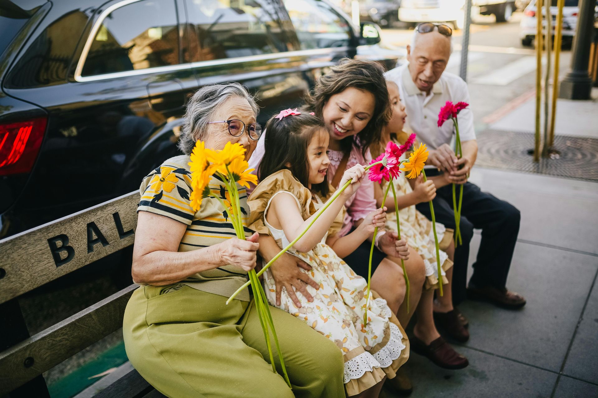 A family is sitting on a bench with flowers in front of a car.