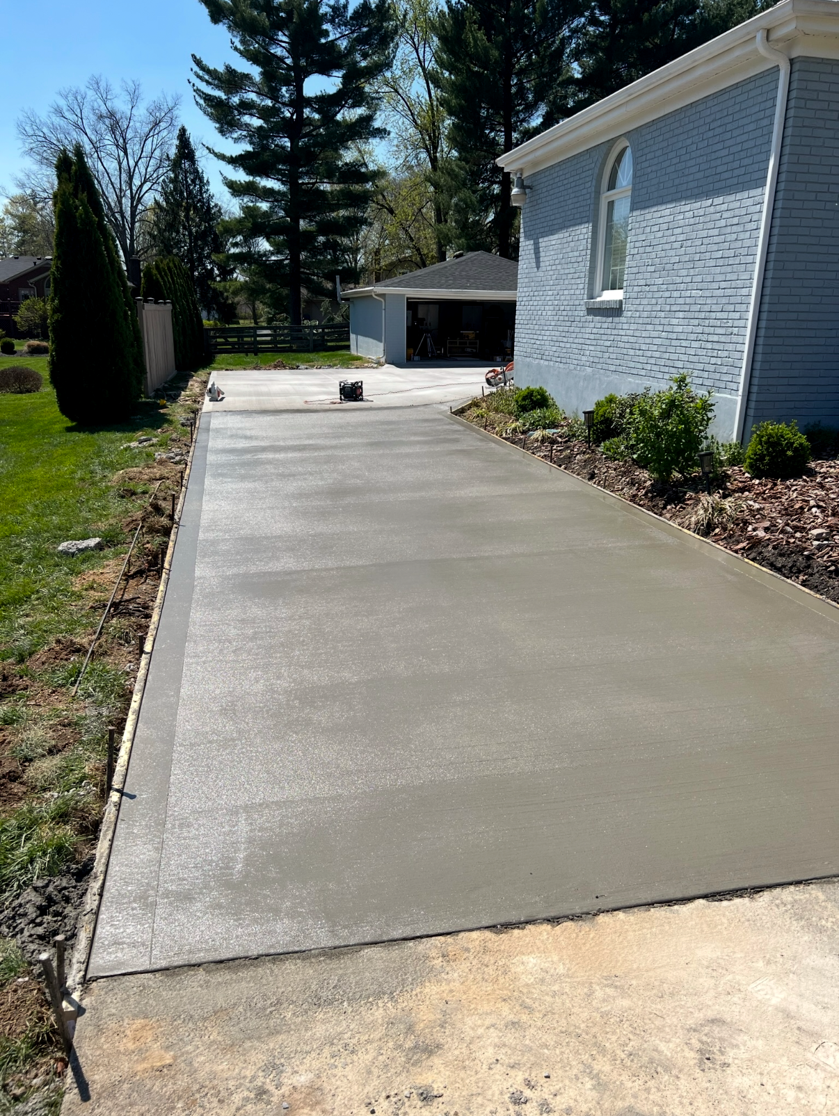 Freshly poured concrete driveway beside a light blue building, green grass, and trees.