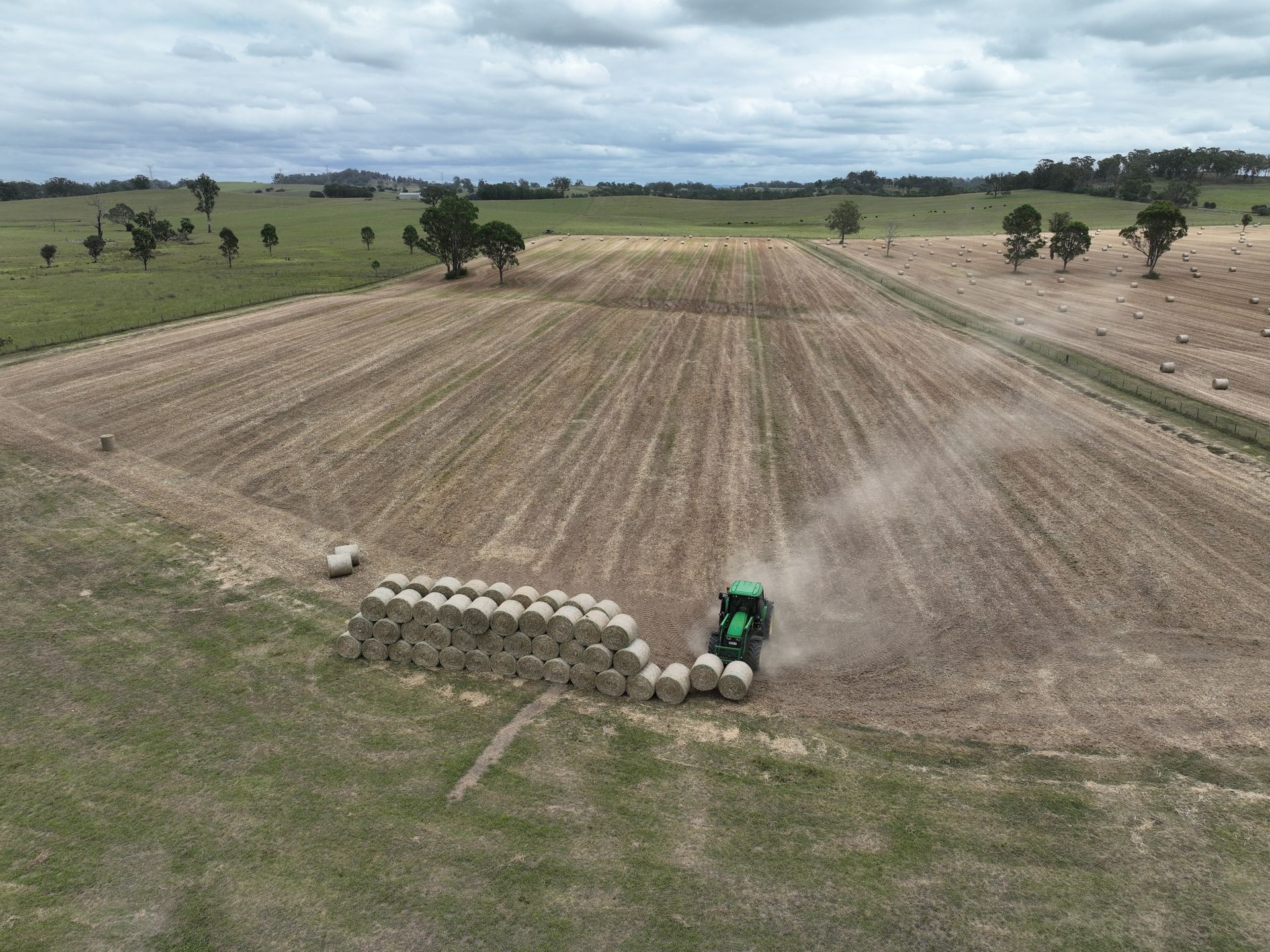 A tractor stacks hay bales in a field. The scene is set on a cloudy day in a rural area with fields and trees.