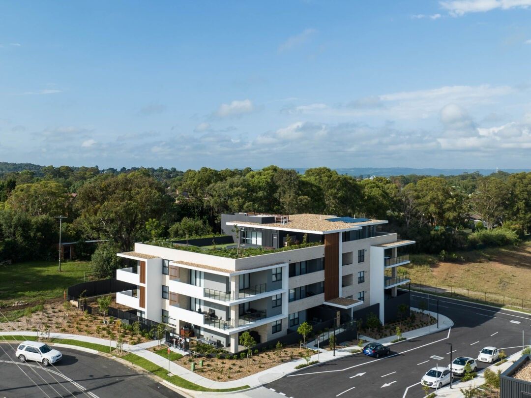 Modern apartment building with white and wood paneling, set on a corner lot surrounded by trees and greenery.