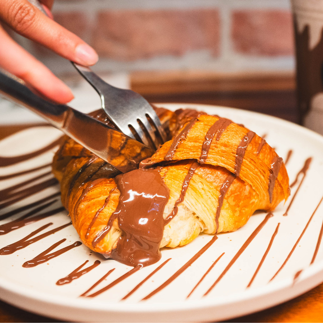 A croissant drizzled with chocolate is being cut open on a plate with chocolate drizzles. Someone is holding a fork and knife.