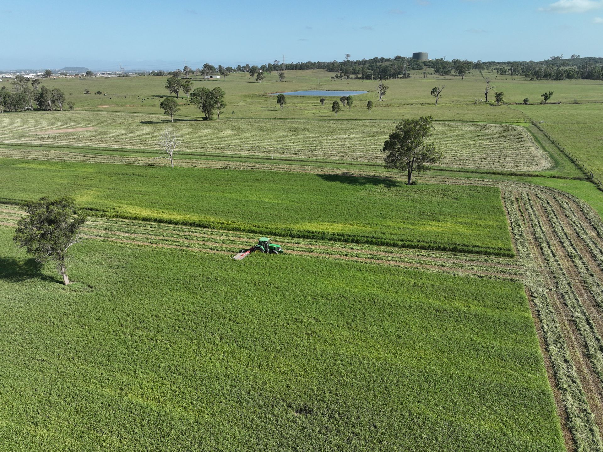Aerial view of a green tractor cutting a path through a field of crops on a sunny day. Fields and trees are visible in the background.