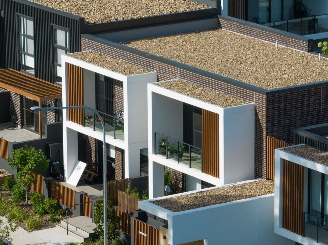 Modern multi-story homes with white walls, brown accents, and flat brown roofs. Balconies have glass railings.
