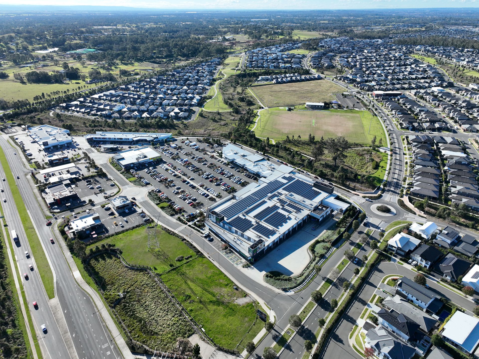 Aerial view of a modern shopping center with a large parking lot and surrounding suburban houses. A highway runs alongside the shopping center.
