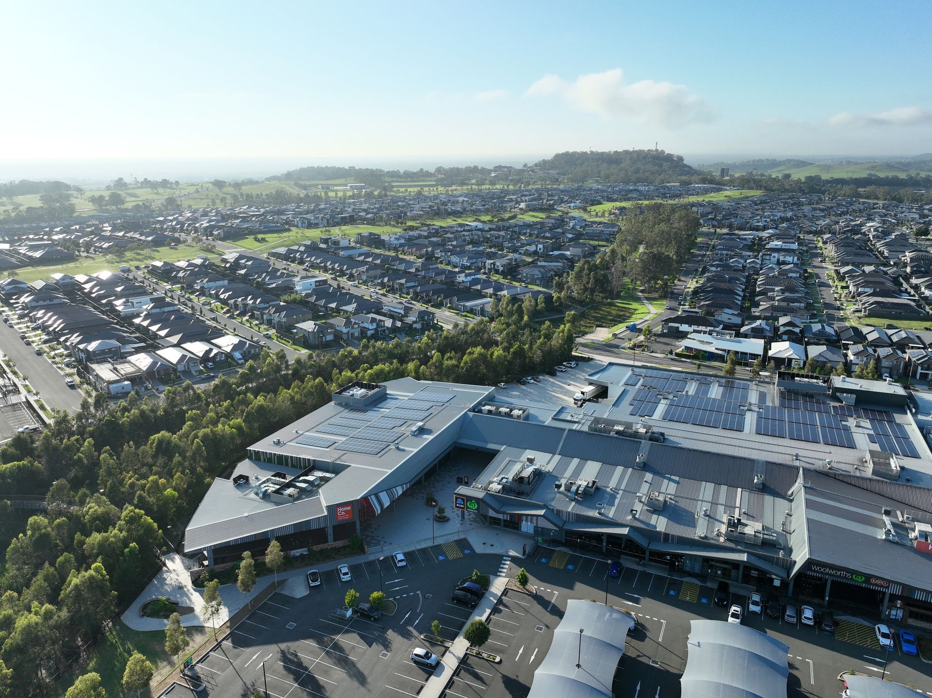 Aerial view of a modern shopping center with parking, surrounded by residential houses and green spaces under a clear sky.
