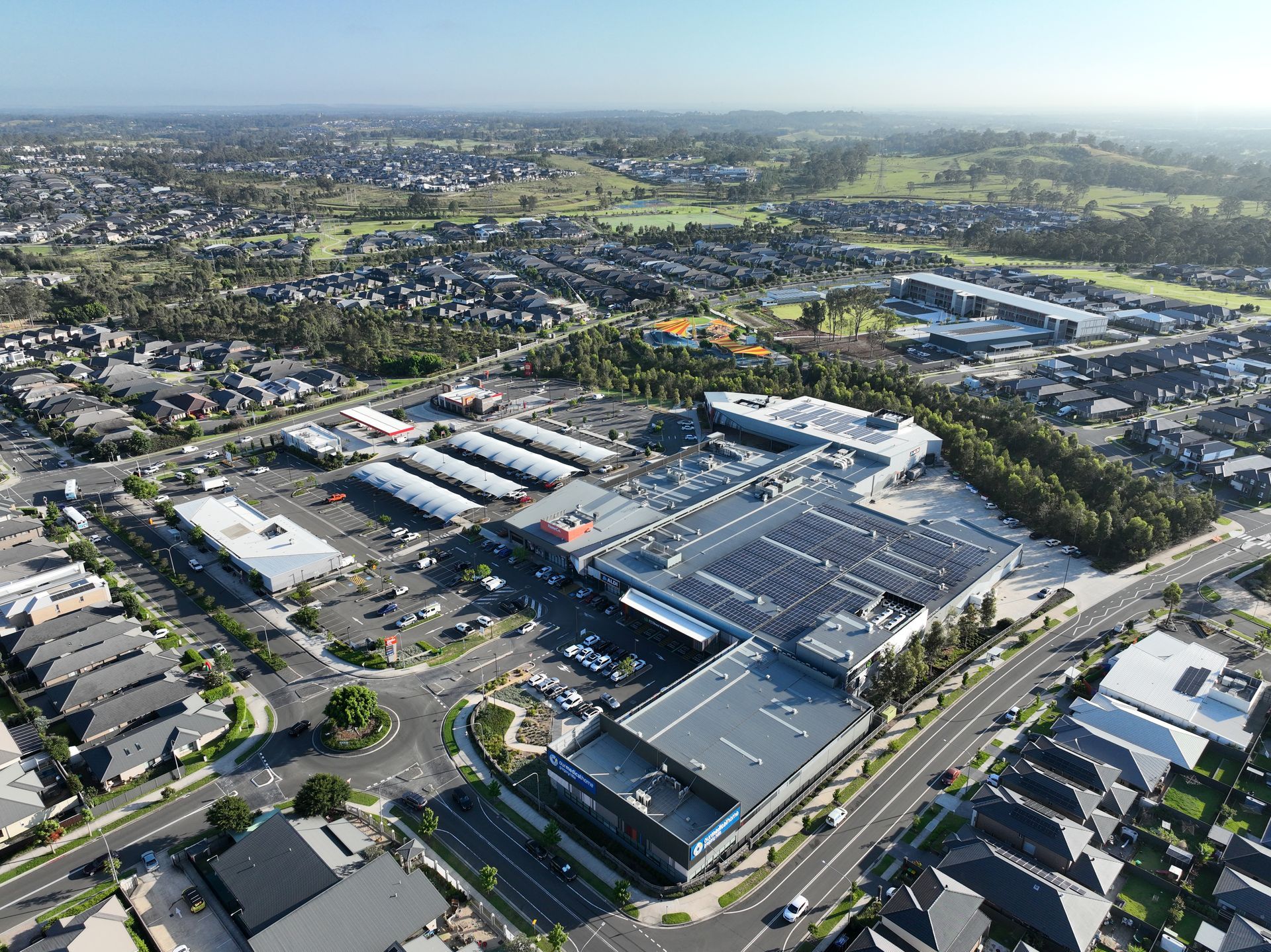 Aerial view of a large shopping center surrounded by residential areas and green space. Sunny, daytime scene.