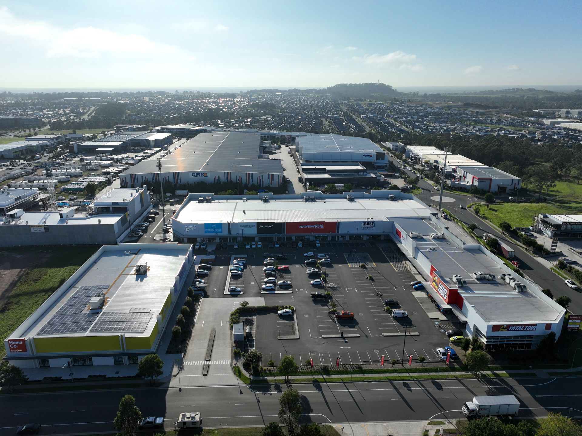 Aerial view of a shopping center and industrial buildings with a large parking lot. The sky is bright.