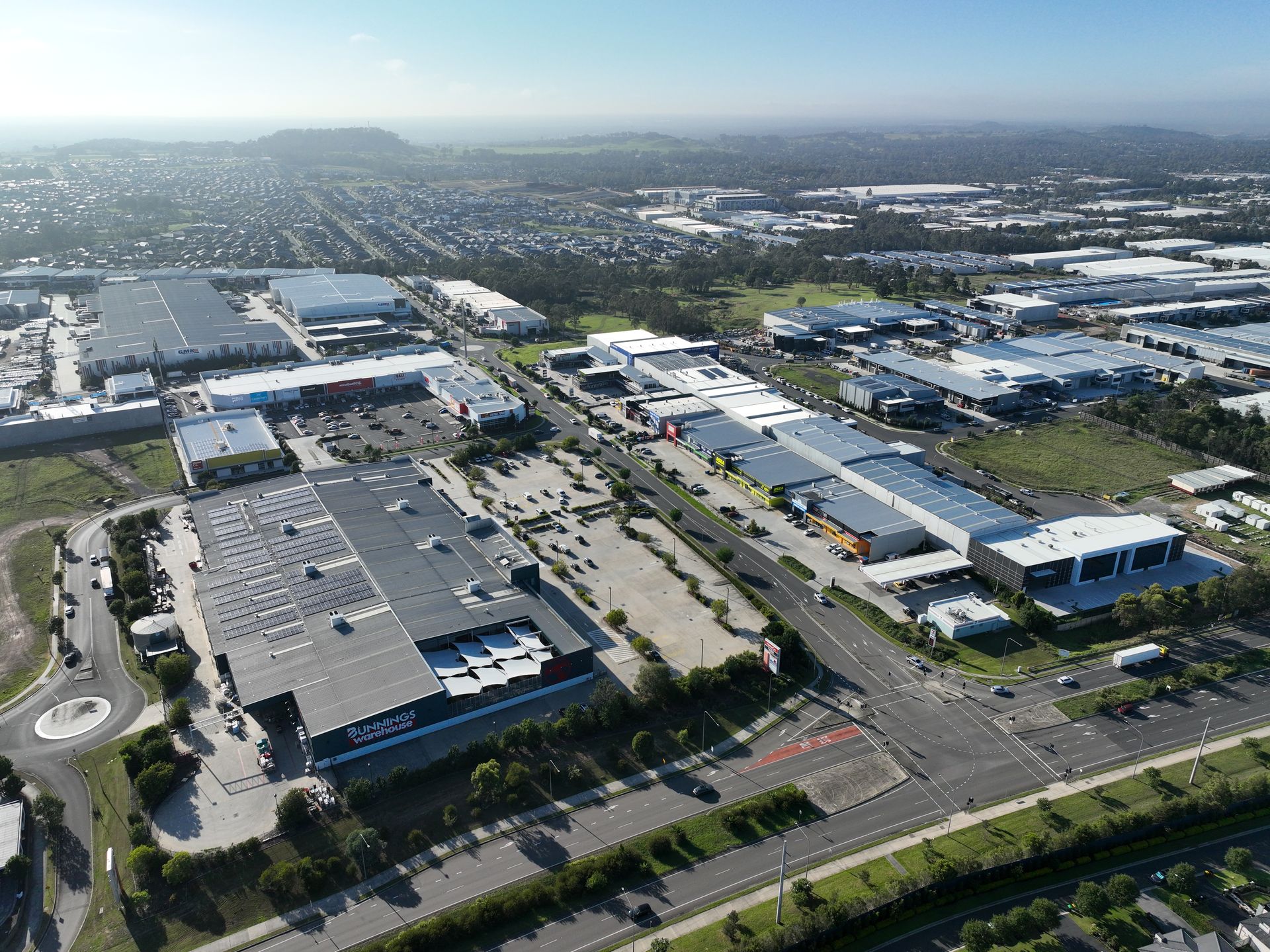 Aerial view of an industrial park featuring large warehouses, retail buildings, and parking lots under a bright sky.