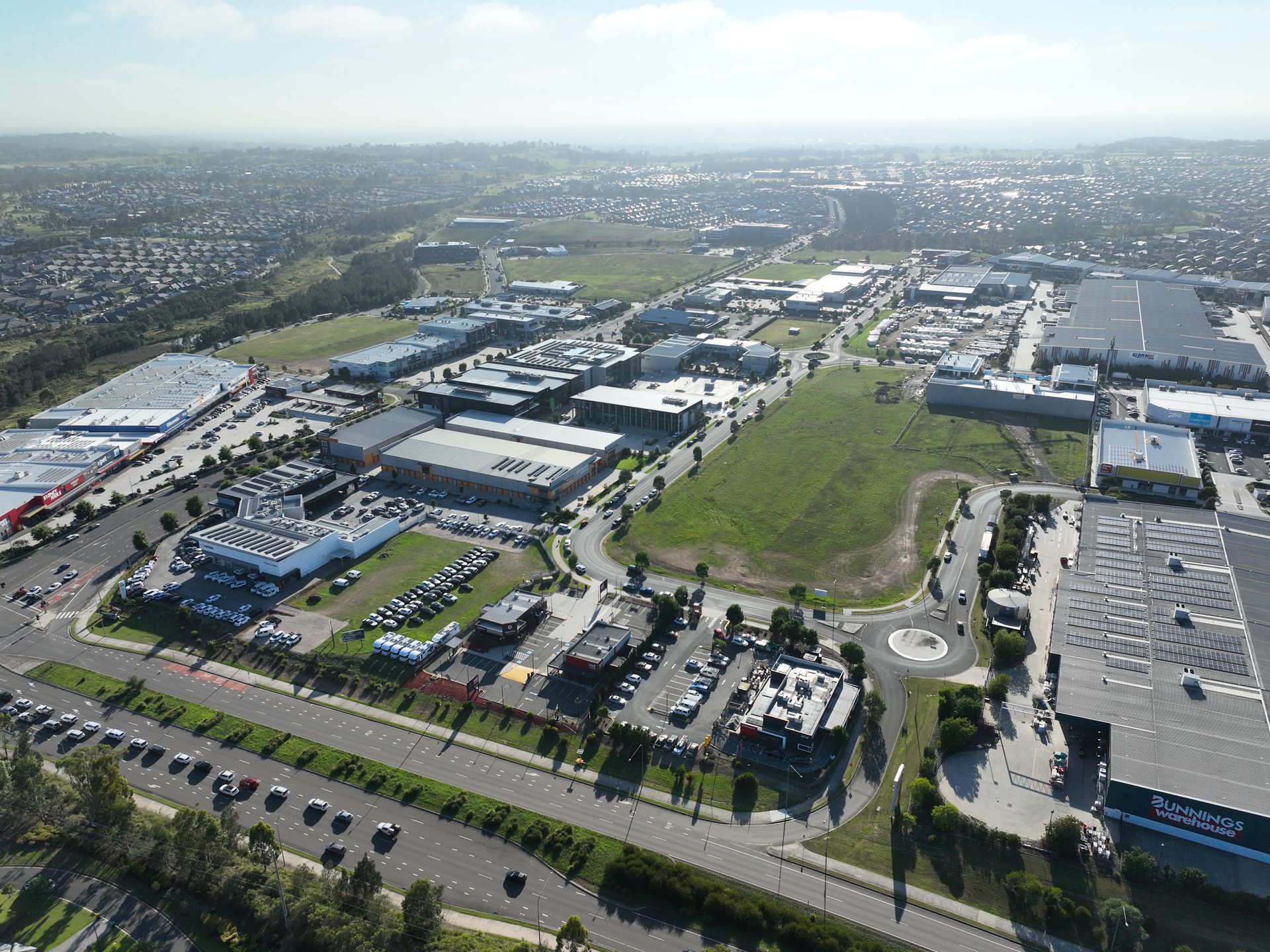 Aerial view of a commercial area with large buildings, parking lots, and a roundabout. Some green space is interspersed.