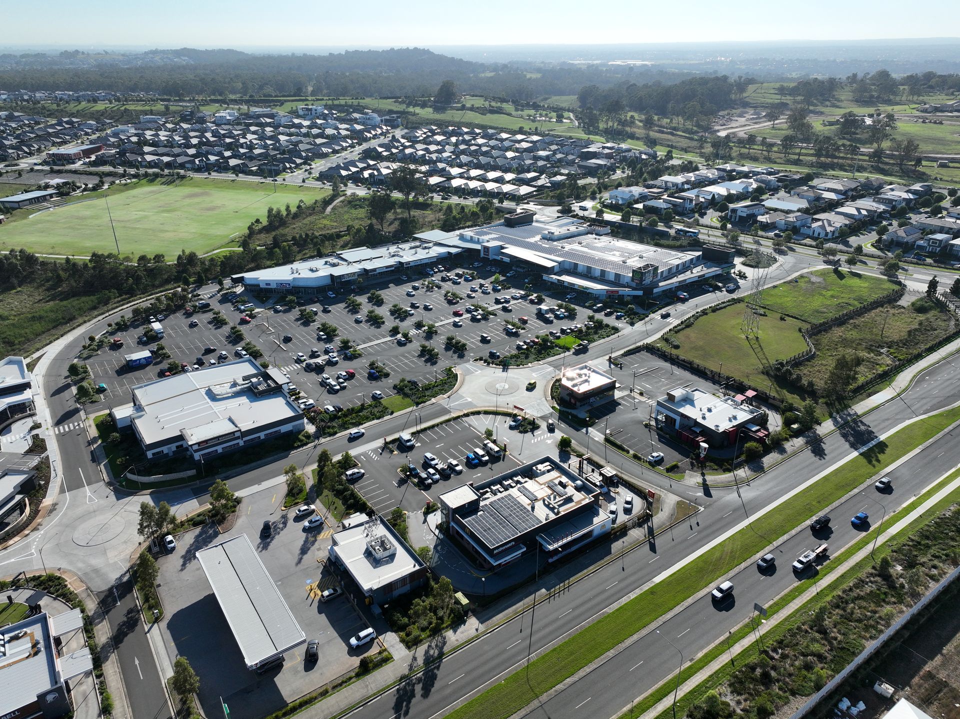 Aerial view of a shopping center with a large parking lot, surrounding buildings, and a road in the foreground. A grassy field and suburban housing are in the background.