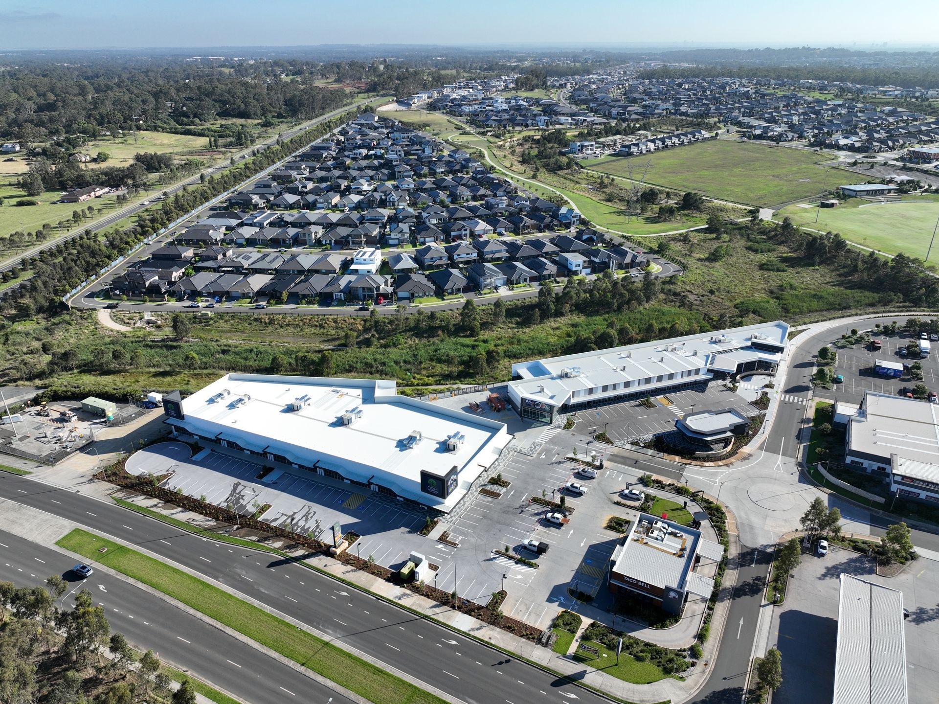Aerial view of a suburban shopping center with a large housing development in the background. The sky is clear, the landscaping is lush.