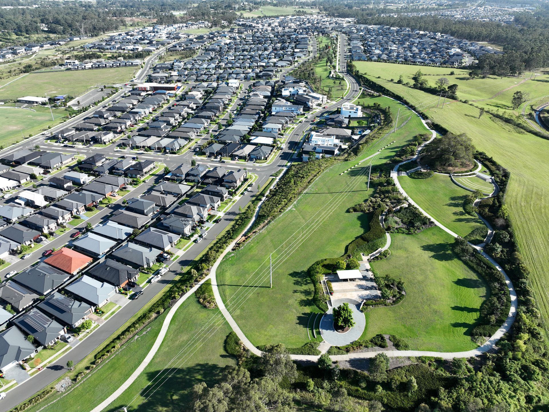 Aerial view of a suburban neighborhood bordering a green park with walking paths. Houses have dark roofs; the park is green.