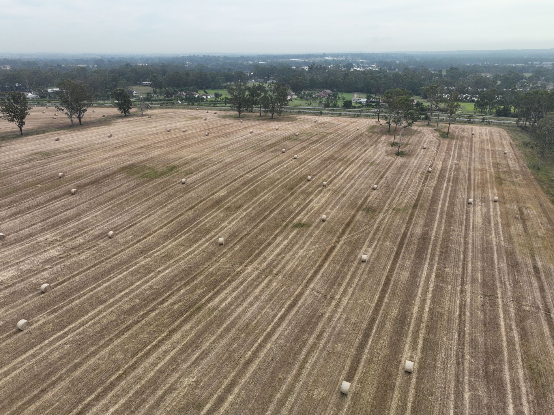 Aerial view of a harvested field with hay bales scattered across it. Trees and a town are visible in the background under a cloudy sky.