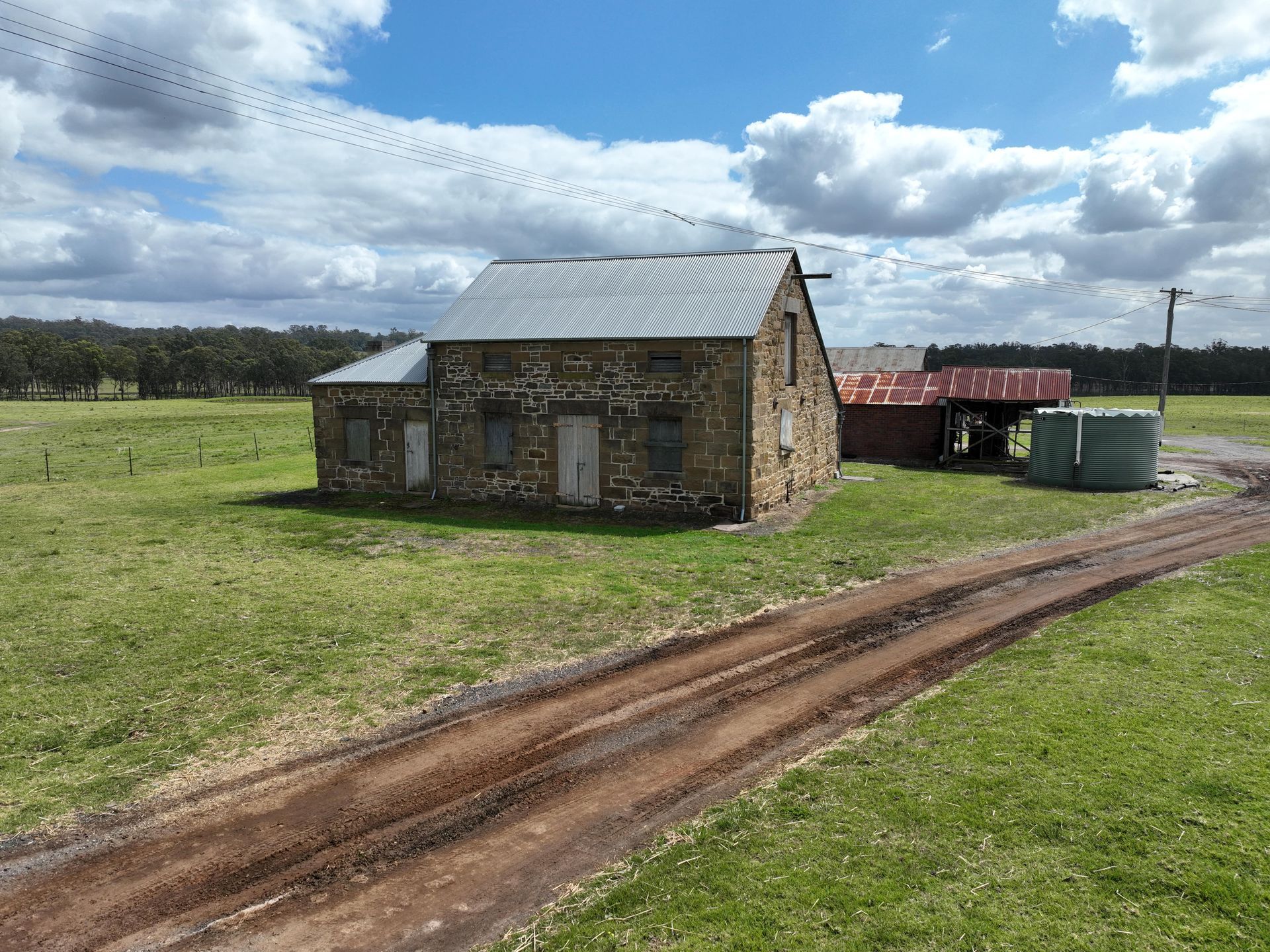 Old stone house with a corrugated metal roof sits on a grassy field. A dirt track leads to it under a cloudy blue sky.
