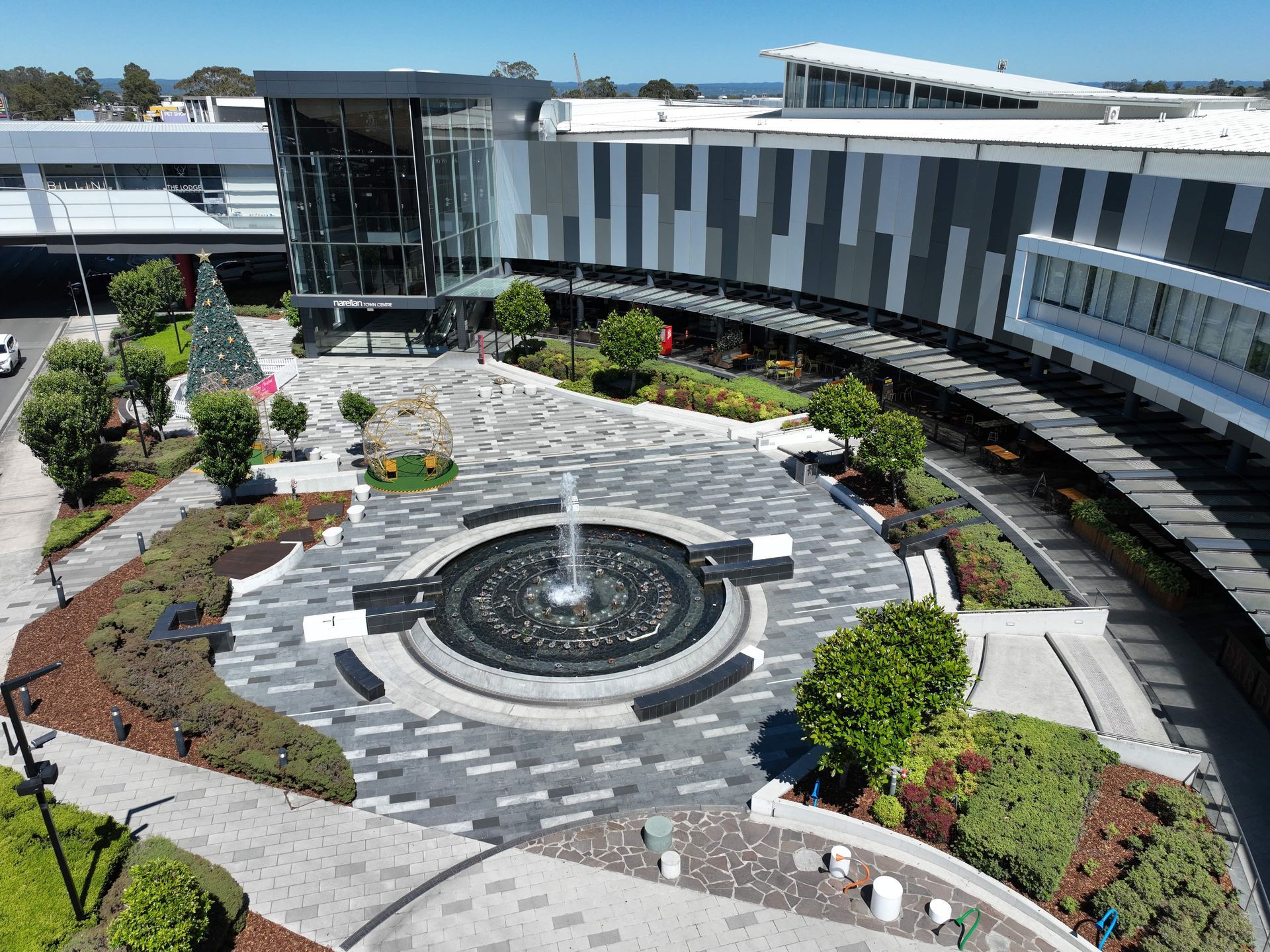 Outdoor plaza with fountain, trees, and modern buildings in an urban setting. Grey and black paving, with green landscaping and a blue sky.