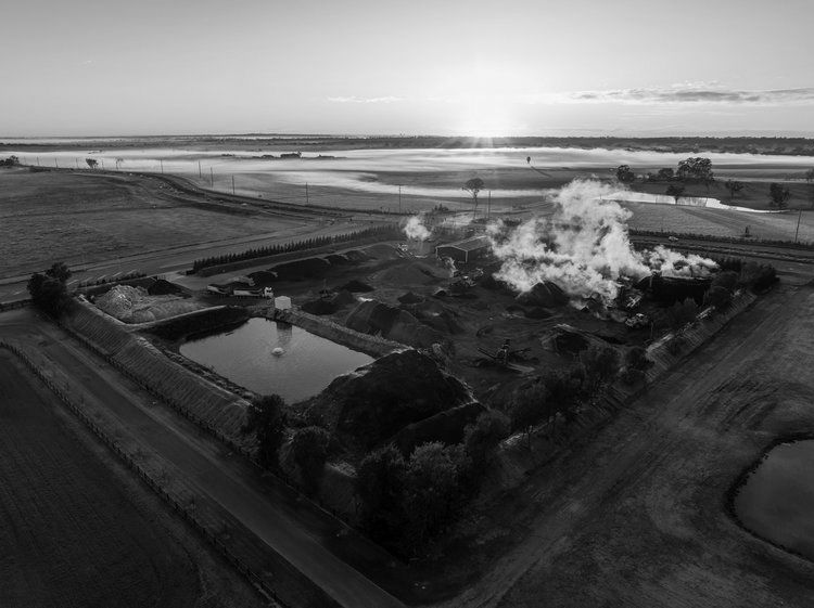 Black and white aerial view of a construction site with smoke rising, a water body, and surrounding fields at dawn.