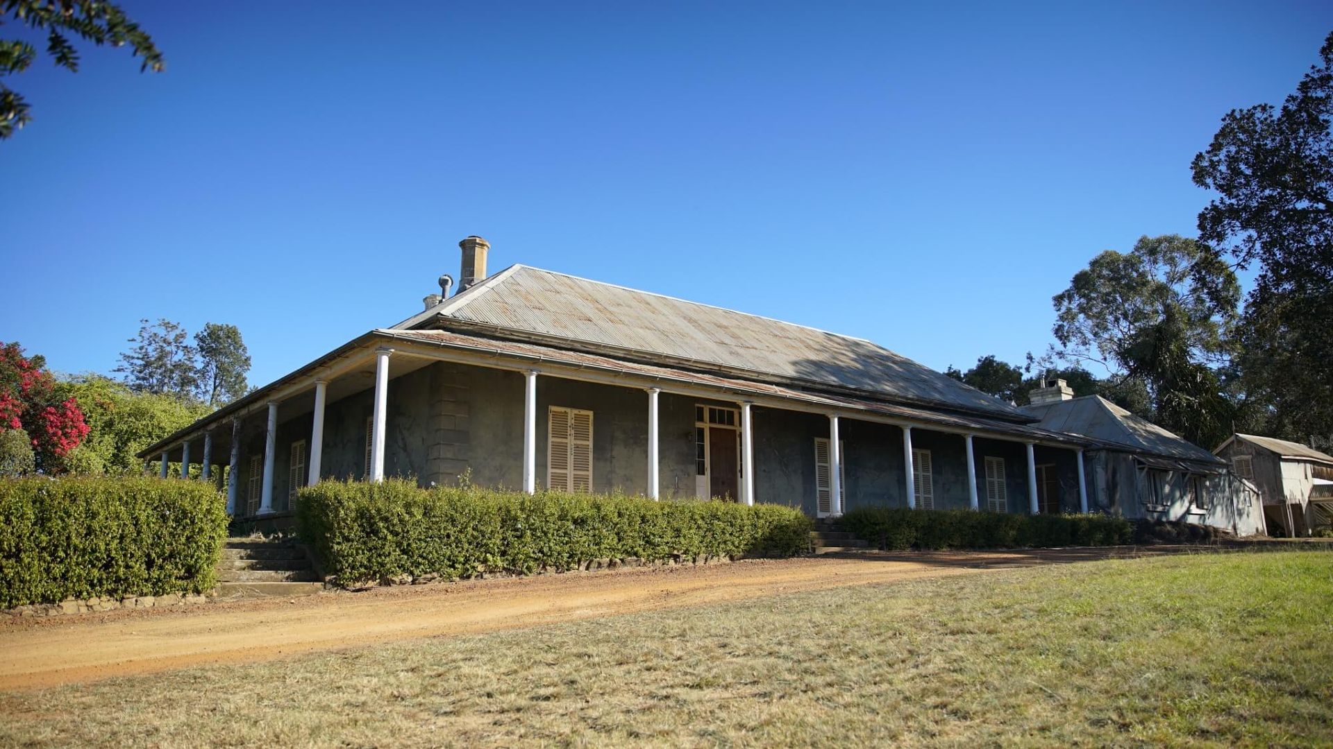 Stone farmhouse with a long veranda under a clear blue sky. A low hedge lines the front; a dirt path and grass lead to the building.