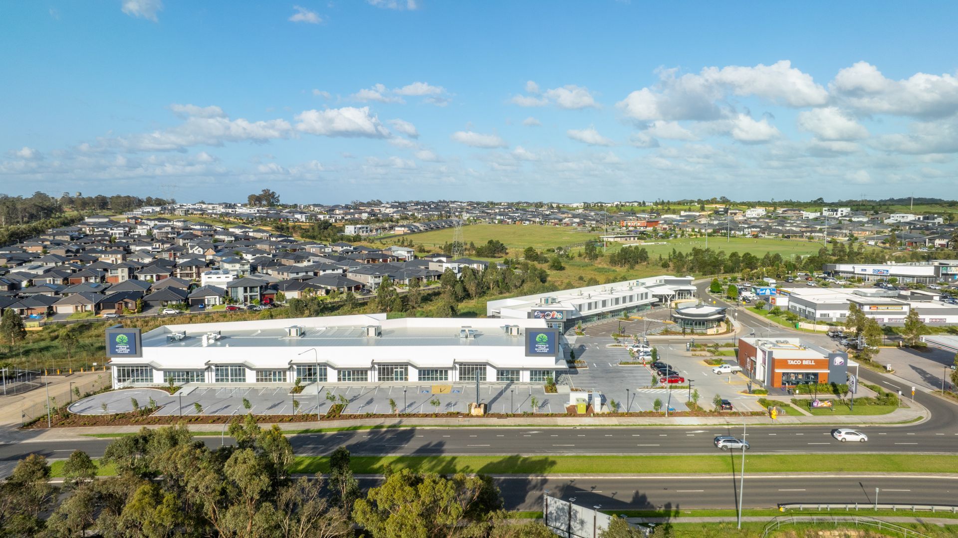 Aerial view of a commercial strip with a long, low building, a McDonald's, and suburban houses under a blue sky.