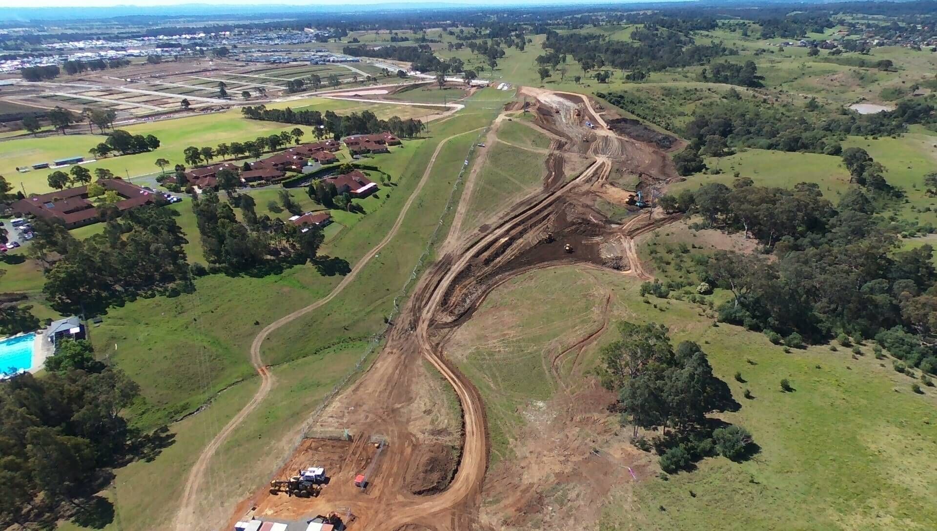 Aerial view of a construction site on a grassy hillside, with heavy machinery visible and dirt paths carved into the landscape.